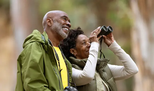 Couple in woods watching their neighbors thru a pair of binoculars and smiling for obvious reasons